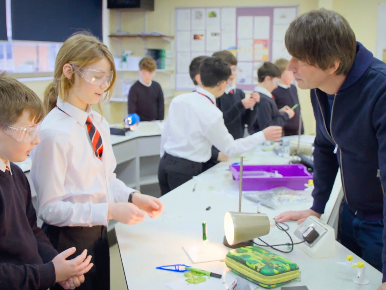 Two children in school uniforms and safety goggles speaking to Brian Cox